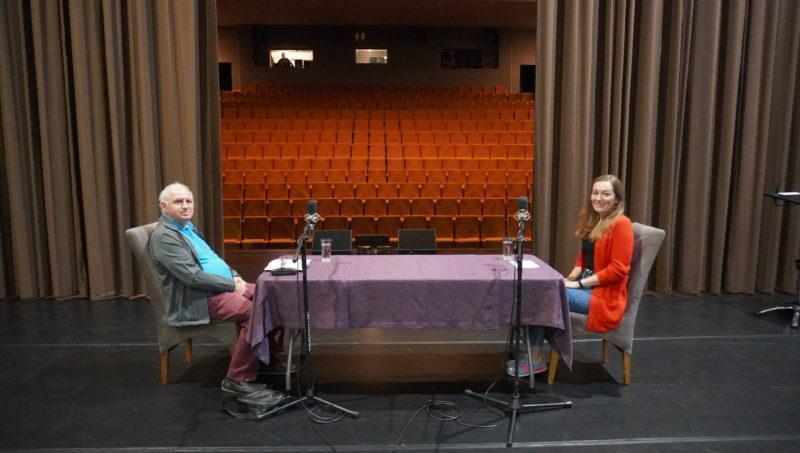 kobieta i mężczyzna siedzący przy stole. Scena w teatrze, w tle pusta widownia, na pierwszym planie dwa mikrofony