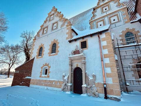 Kościół Świętej Trójcy w Żórawinie Widok kościoła z zewnątrz, nieduże drzwi łukowe zdobione niewielkimi kolumienkami po jednej i drugiej stronie. Elewacja biała zdobiona w kolorze beżowym na szczytach i wokół okien