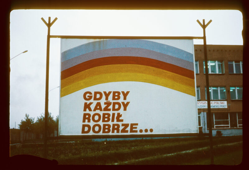 Władysław Hasior o tytule "Notatnik fotograficzny" z serii "Poezja przydrożna" z lat 1965-1998. W ramie stojącej na dworze, plakat z tęczą, pod nią napis "Gdyby każdy robił dobrze..."