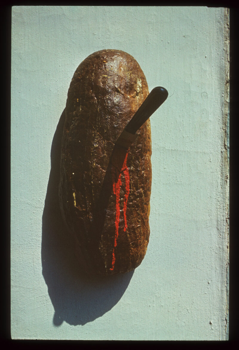 Władysław Hasior "Notatnik fotograficzny" z lat 1965-1998, z serii "Miniatury, chleb, maszyna". Przedstawia bochenek chleba od góry, z wbitym nożem, z rany cieknie krew.