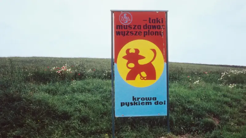 Władysław Hasior, „Notatnik fotograficzny”, z serii "Poezja przydrożna". Na łące, w ramie stoi plakat, z grafiką krowy w kole. Na górze napis: "-łąki muszą dawać wyższe plony". Pod ilustracją "krowa pyskiem doi".