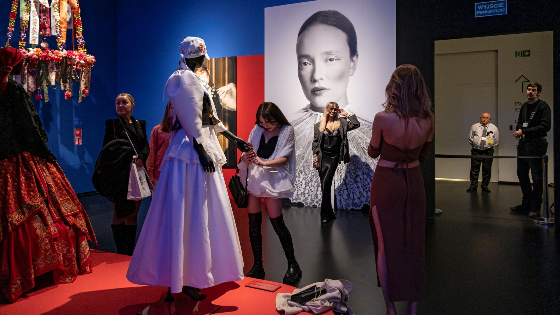 Od lewej: Manekiny ubrane w stroje ludowe, po środku zwiedzający robiący zdjęcia, po prawej pracownicy pilnujący przestrzeni ekspozycji