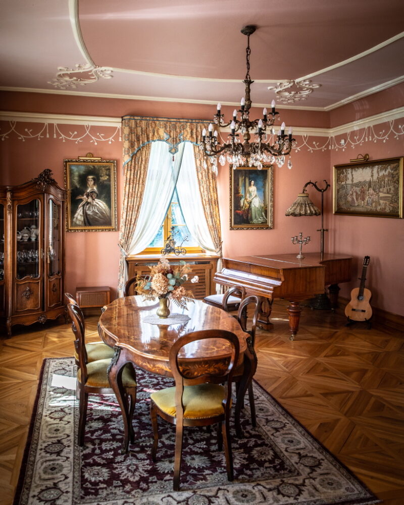 maslow_V9A1587_1024 Jeden z pokoi dworu, w stylu barokowym. Na środku na dywaniku stoi stół z krzesłami, na ukos po prawo stoi fortepian.