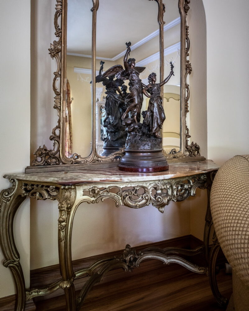 maslow_V9A2133_1024 Zdjęcie toaletki w stylu barokowym z ozdobnym lustrem. Na niej stoi statuetka dwóch kobiet z motywem starożytnym.