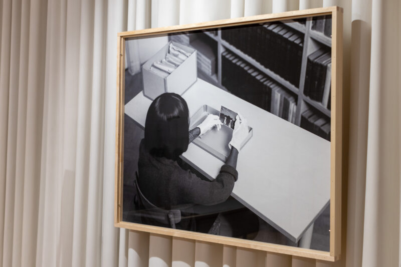 Fotografia Witka Orskiego „Kobieta ze zdjęciem”, wystawa „Wybielanie”. Na fotografii kobieta siedzi przy biurku i patrzy na zdjęcie.