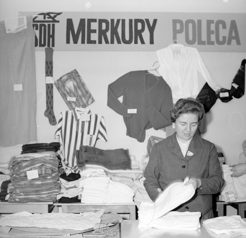PIC_40-4-248-3__OBR SDH "Merkury" Warszawskiej Spółdzielni Spożywców "Społem" przy ulicy Słowackiego w Warszawie od środka. W pokoju siedzi kobieta przy biurku, za nią reklama z napisem "Merkury Poleca". Zdjęcie z 1967 roku.